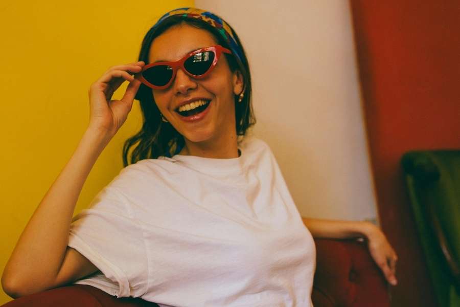 Une femme souriante en t-shirt blanc ajuste ses lunettes de soleil triangle rouges rétro.